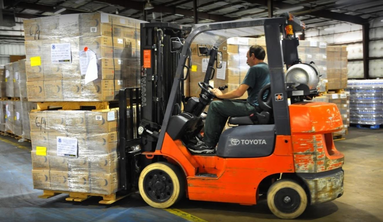 Forklift in warehouse; blue and orange, ready for use, pallets in background.
