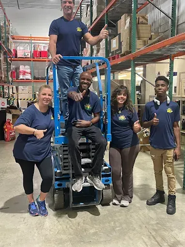 Group of people in matching shirts in a warehouse. Some give thumbs up. One person sits on a lift.
