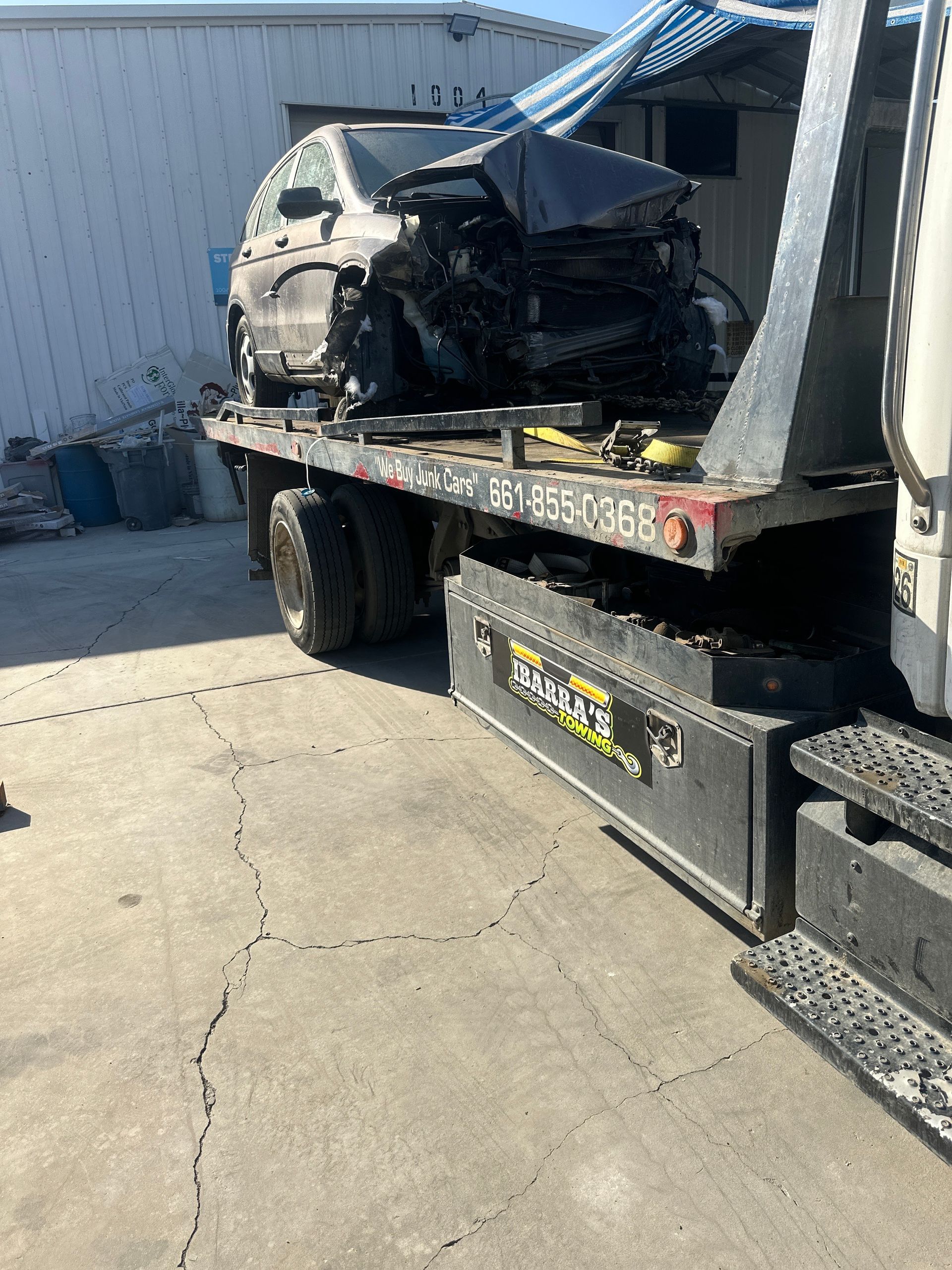 Wrecked car on a tow truck bed; gray vehicle, crumpled front, on concrete outside a building.