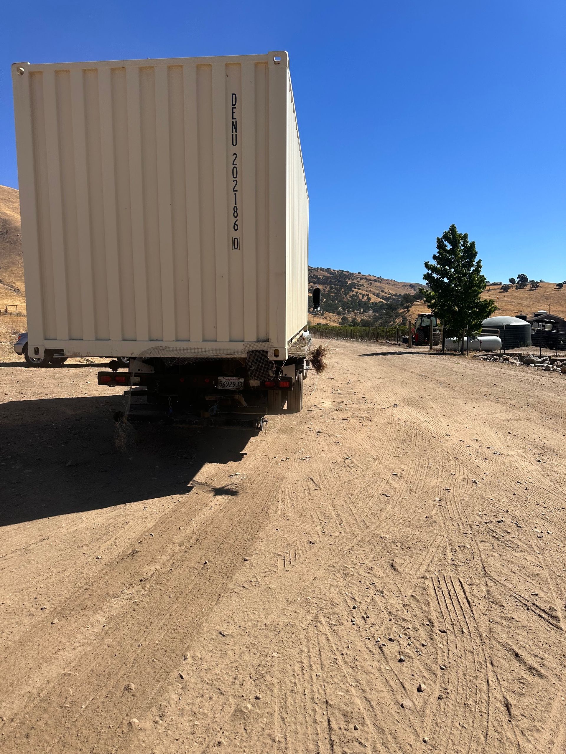 A large cream-colored shipping container sits on a trailer in a dirt field under a blue sky.