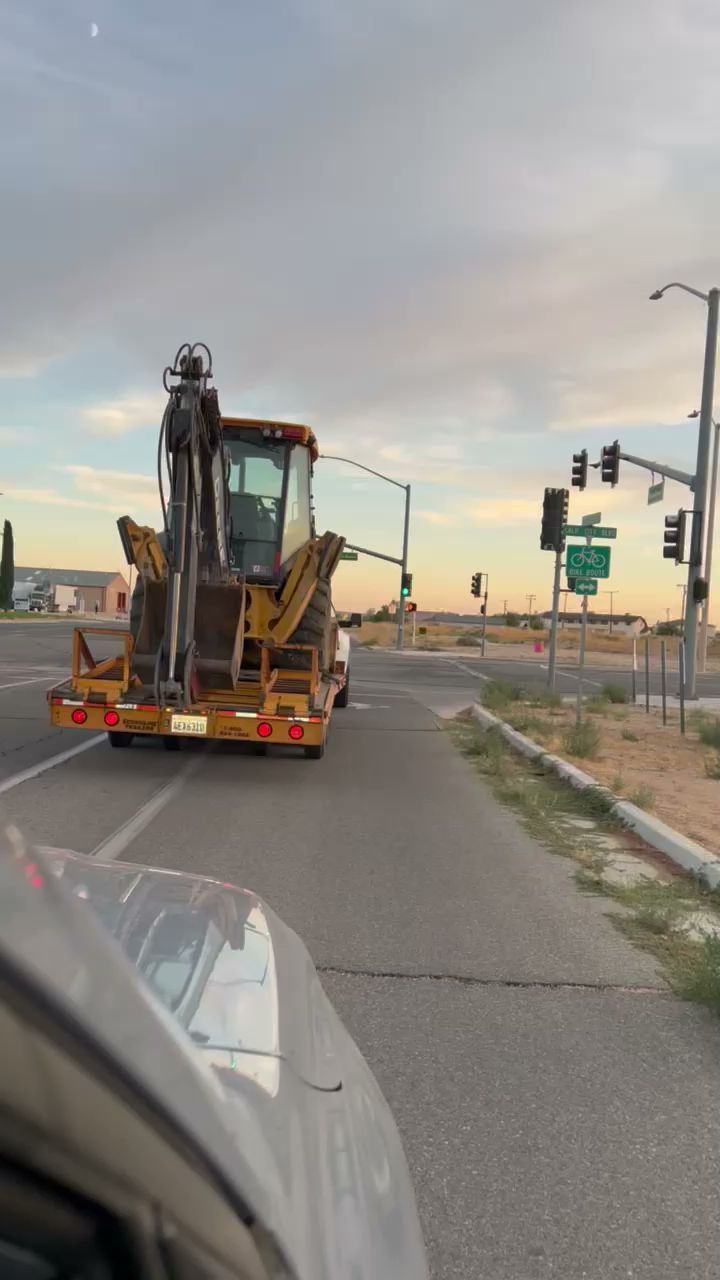 Backhoe on a trailer at an intersection, viewed from inside a car. Dusk sky, traffic lights.