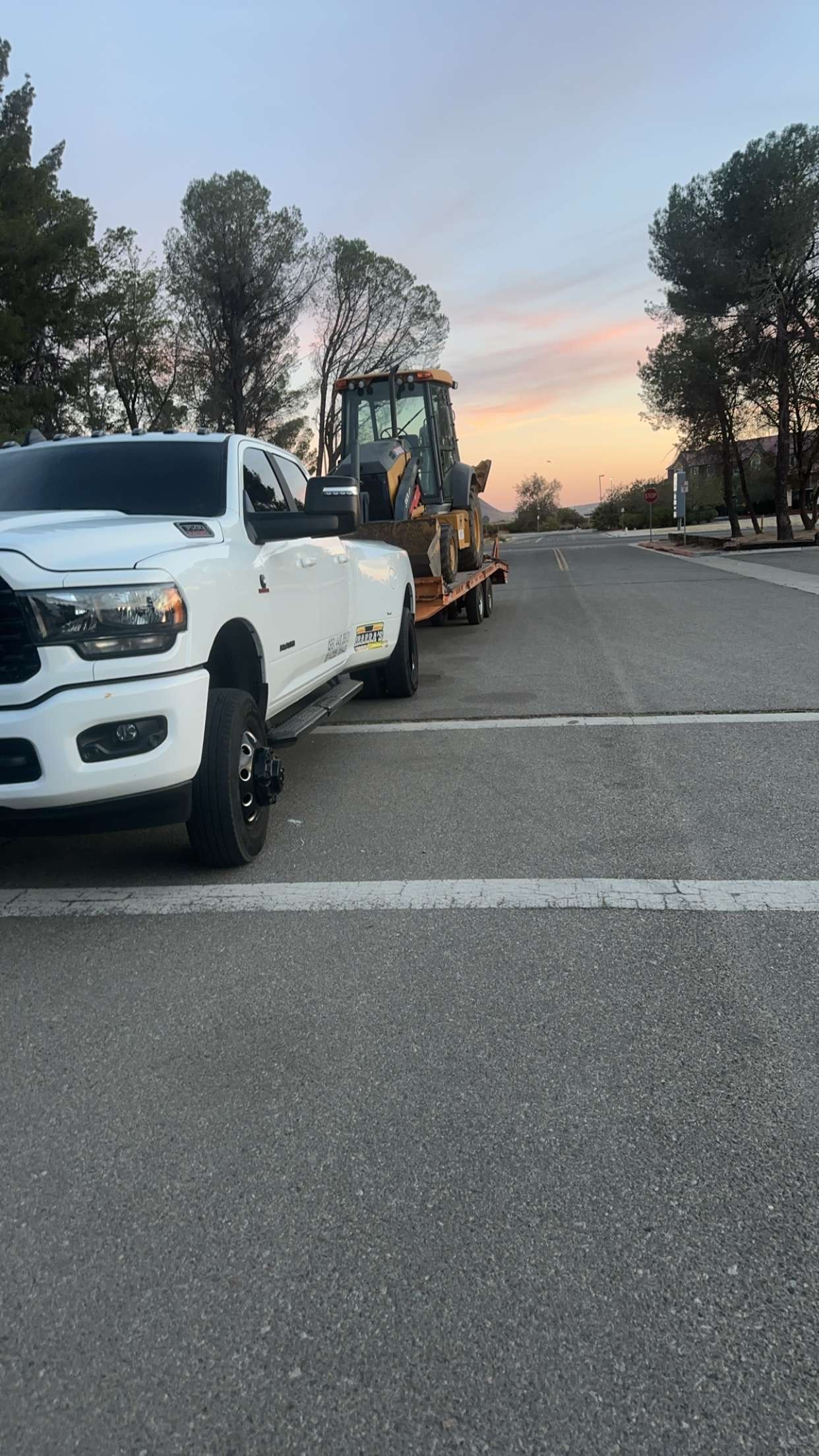White truck towing a yellow construction vehicle on a road, sunset in background.