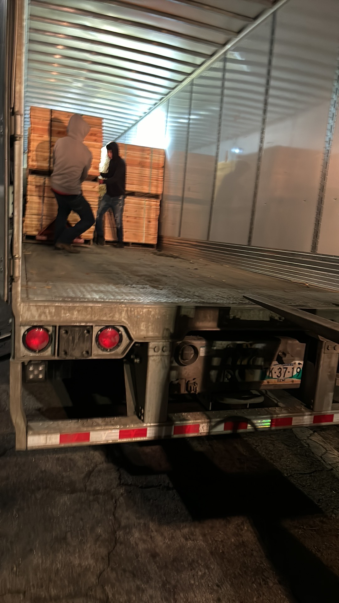 Two men loading wood pallets into an open semi-trailer at night.