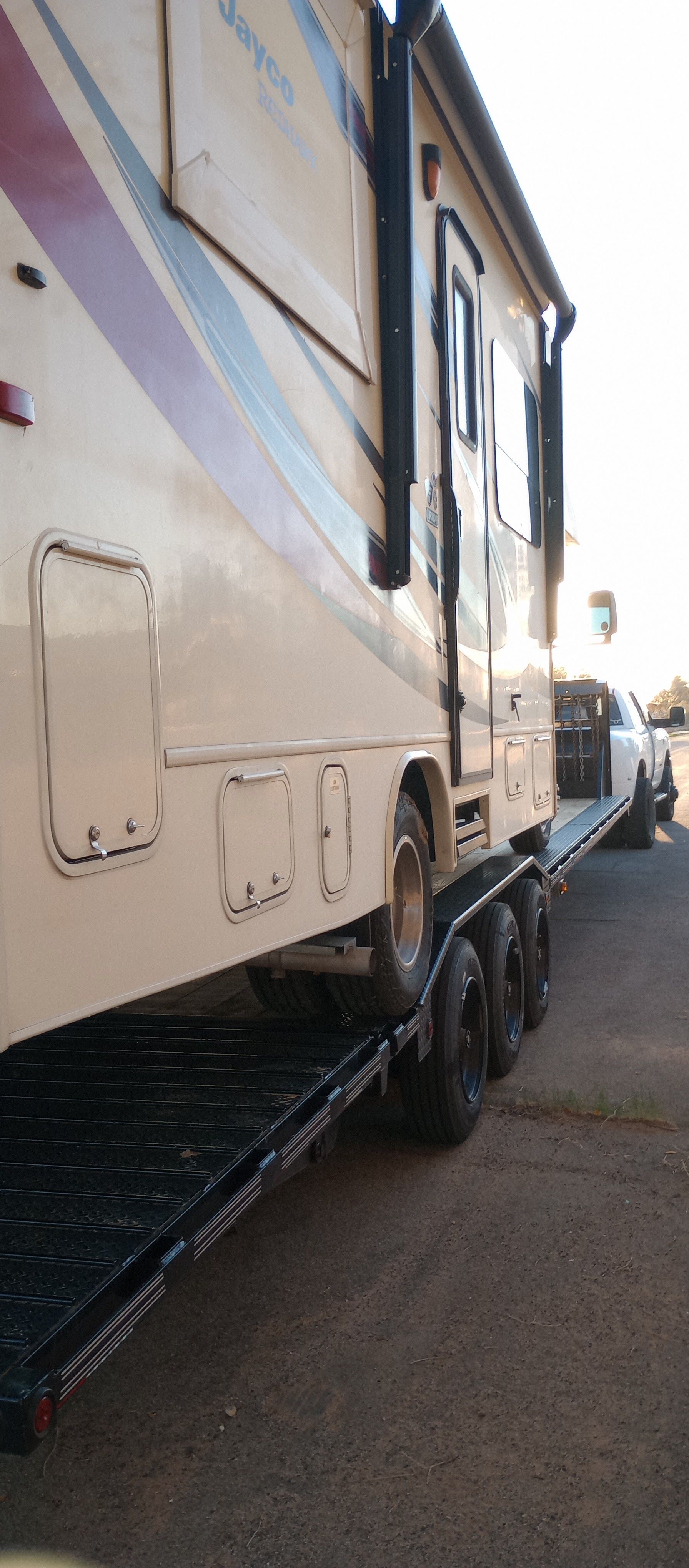 A beige RV is being hauled on a black flatbed trailer by a white truck.