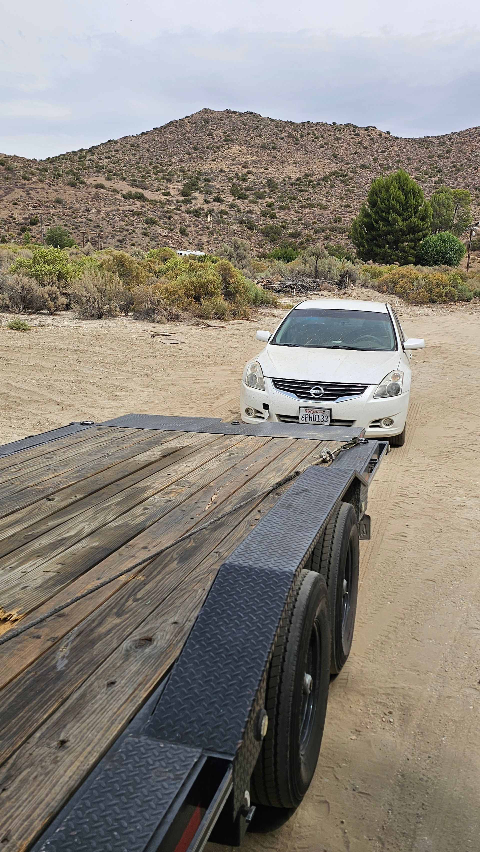 White car loaded on a trailer on a dirt road, with a rocky hillside in the background.
