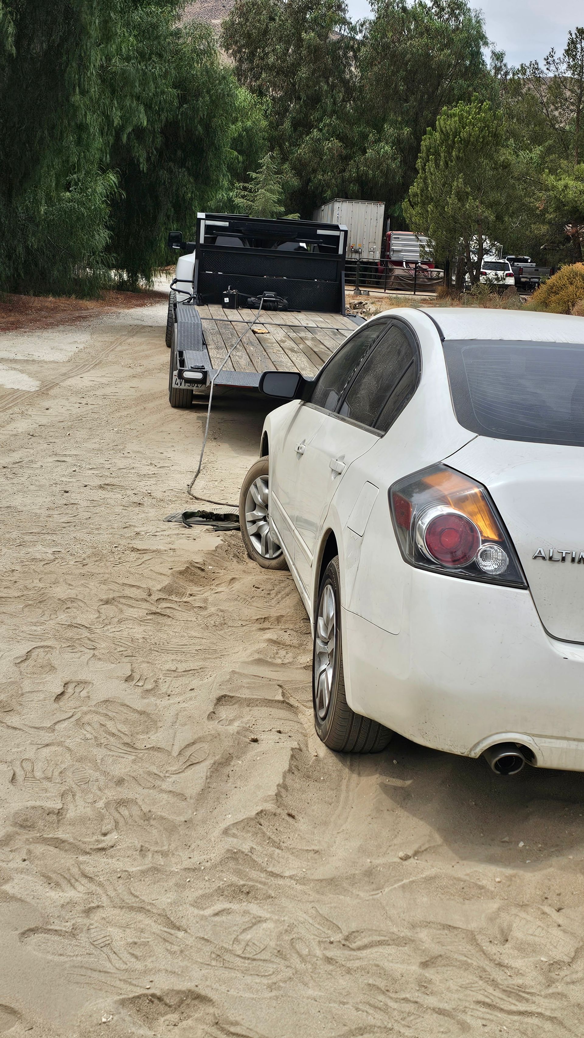 White car being loaded onto a flatbed trailer on a sandy surface, outdoors.