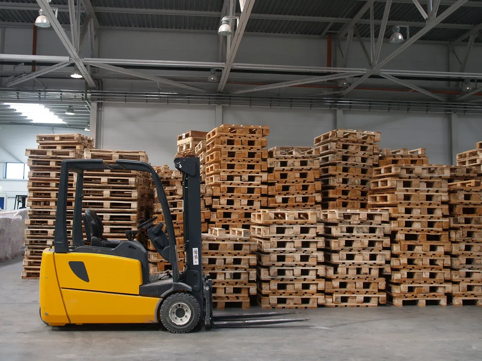 Yellow forklift in a warehouse, in front of a large stack of wooden pallets.
