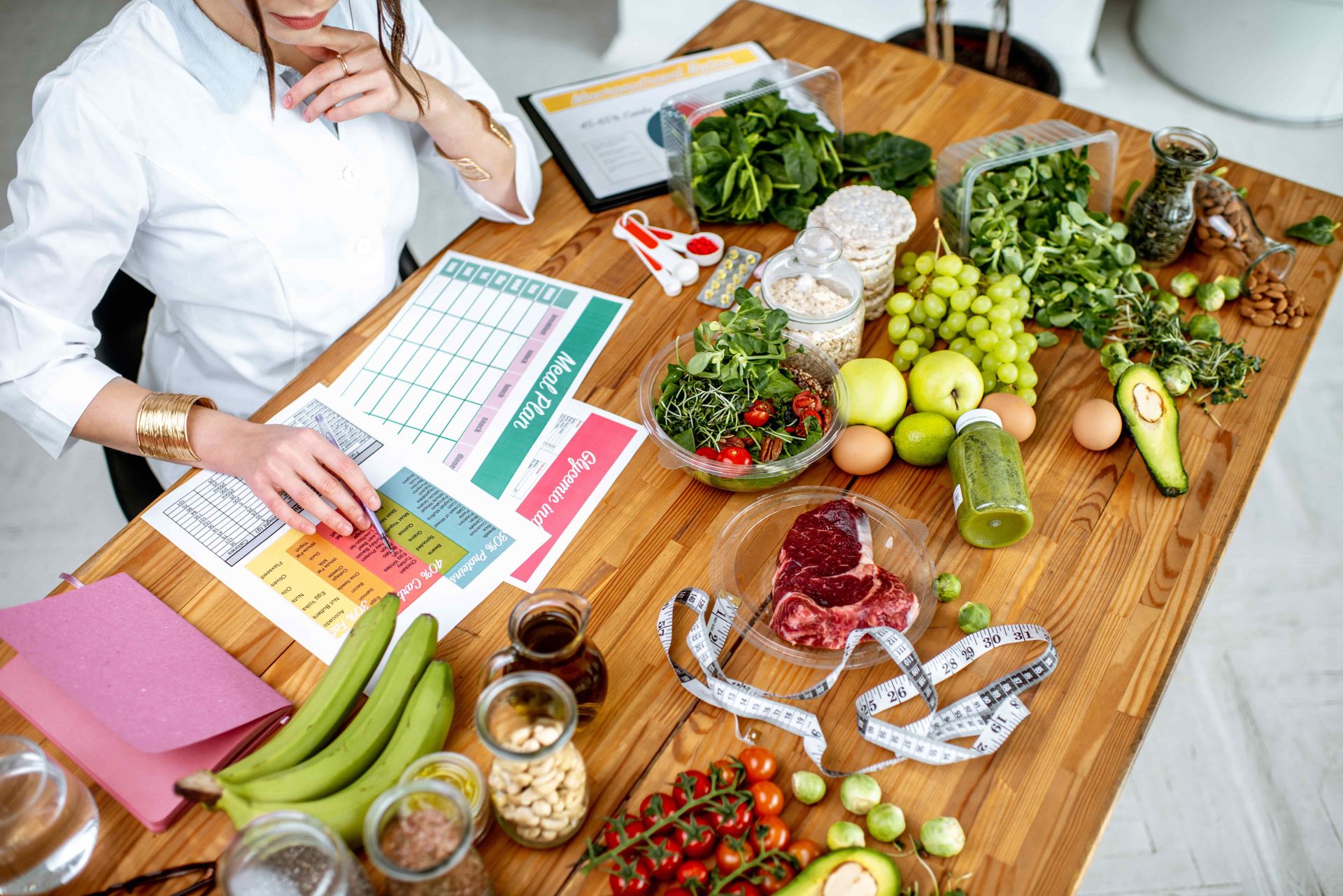 Woman with a diet plan, surrounded by fresh fruits, vegetables, meat, and nuts on a wooden table.