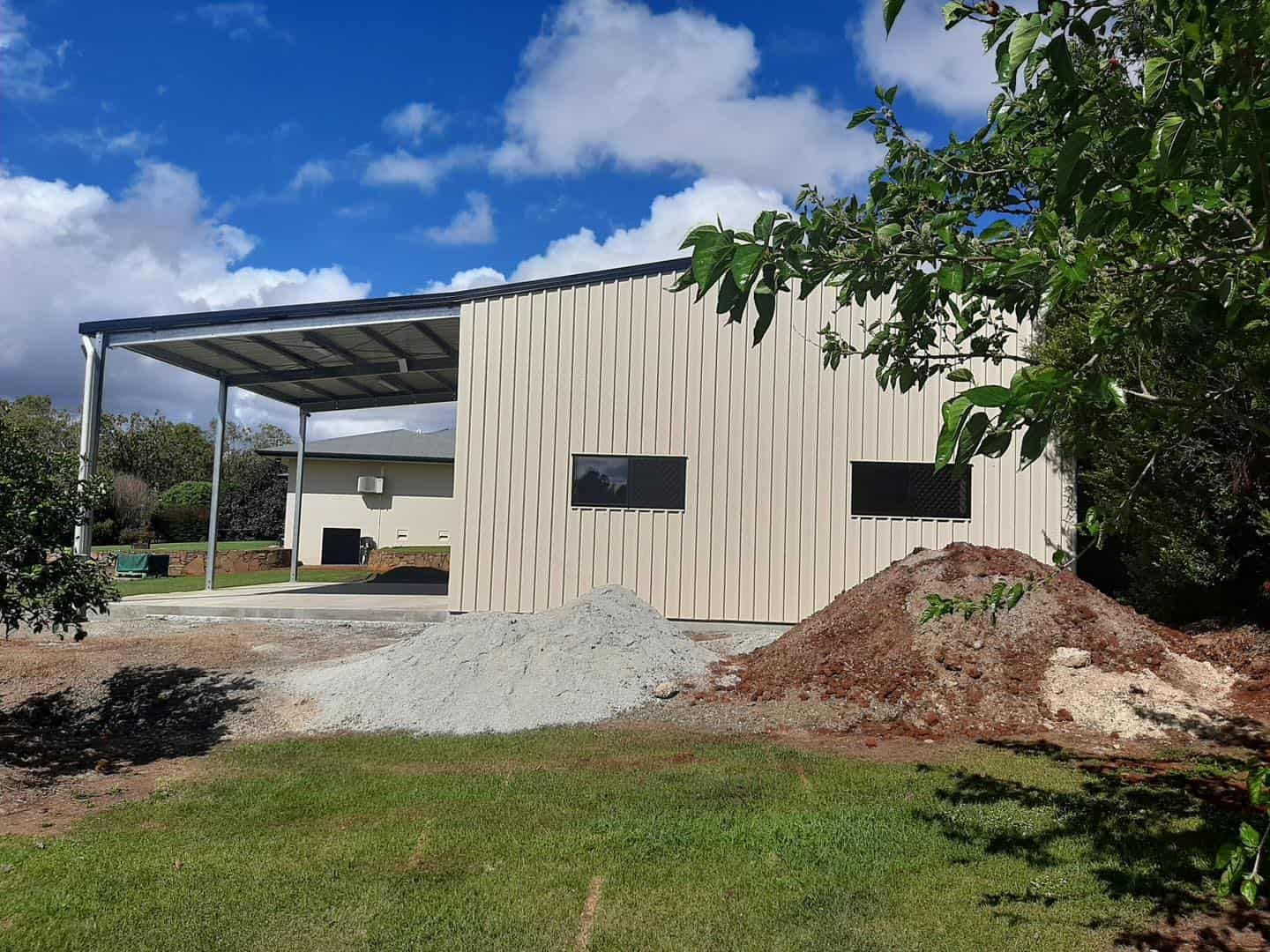 Corrugated Metal Building With a Covered Awning, Gravel Pile, and Green Grass Against a Blue Sky — ShedBoss in Atherton, QLD