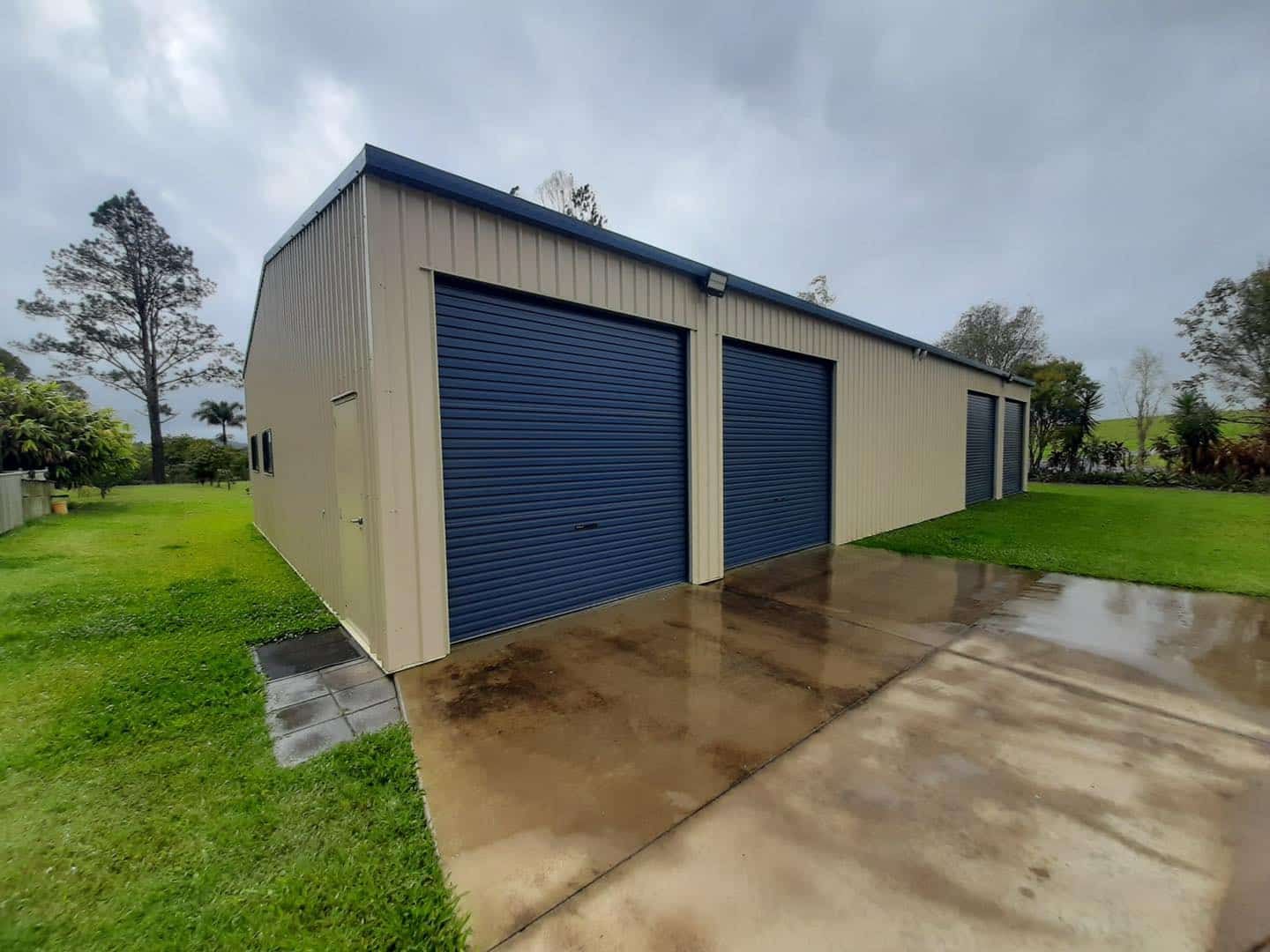 Tan Metal Shed With Three Blue Garage Doors on a Wet Concrete Driveway — ShedBoss in Atherton, QLD