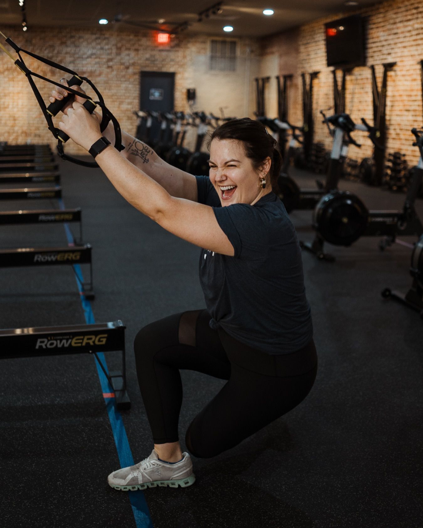 Woman doing a suspension squat in a gym, smiling and holding TRX straps.