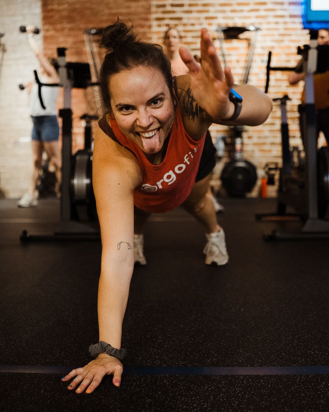 Smiling woman lunges toward the camera in a gym, wearing a red tank top and black workout leggings.