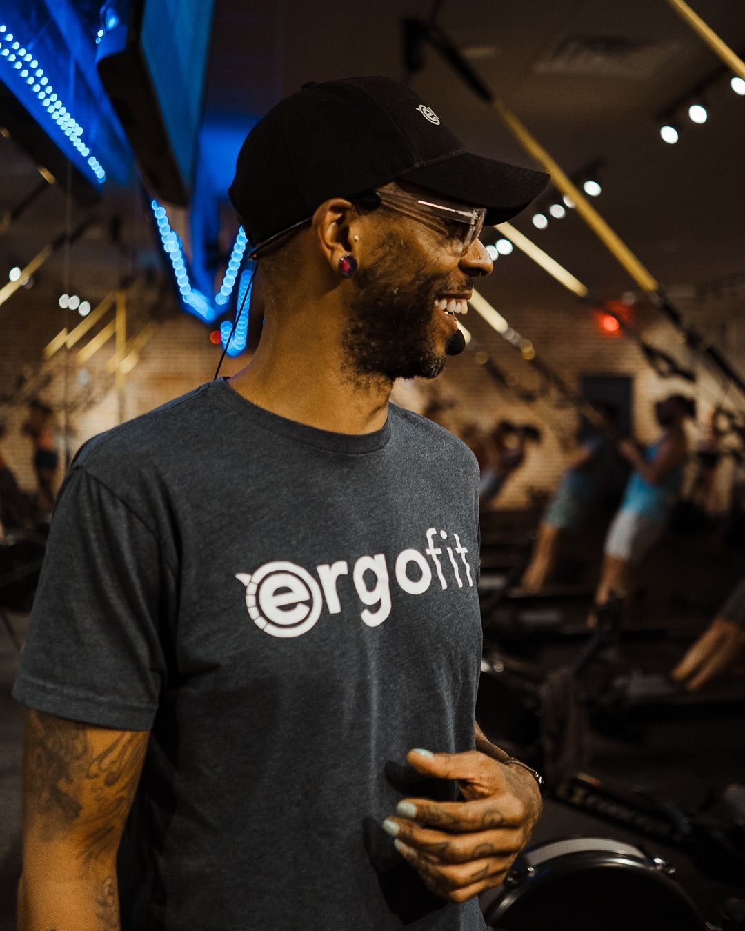 Man in black cap and glasses wearing an Ergofit T-shirt in a gym, smiling and looking left