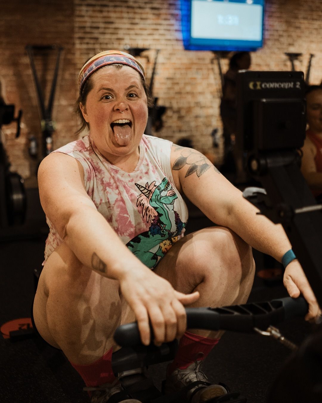 Person rowing on a machine in a gym, smiling and wearing a colorful tank top and headband.