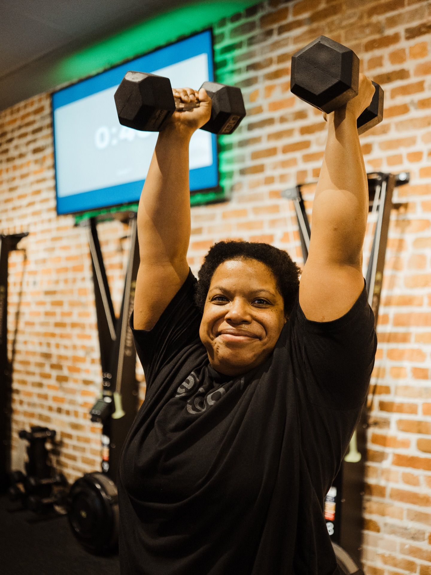 Person lifting dumbbells overhead in a gym, smiling in front of a brick wall and TV screen