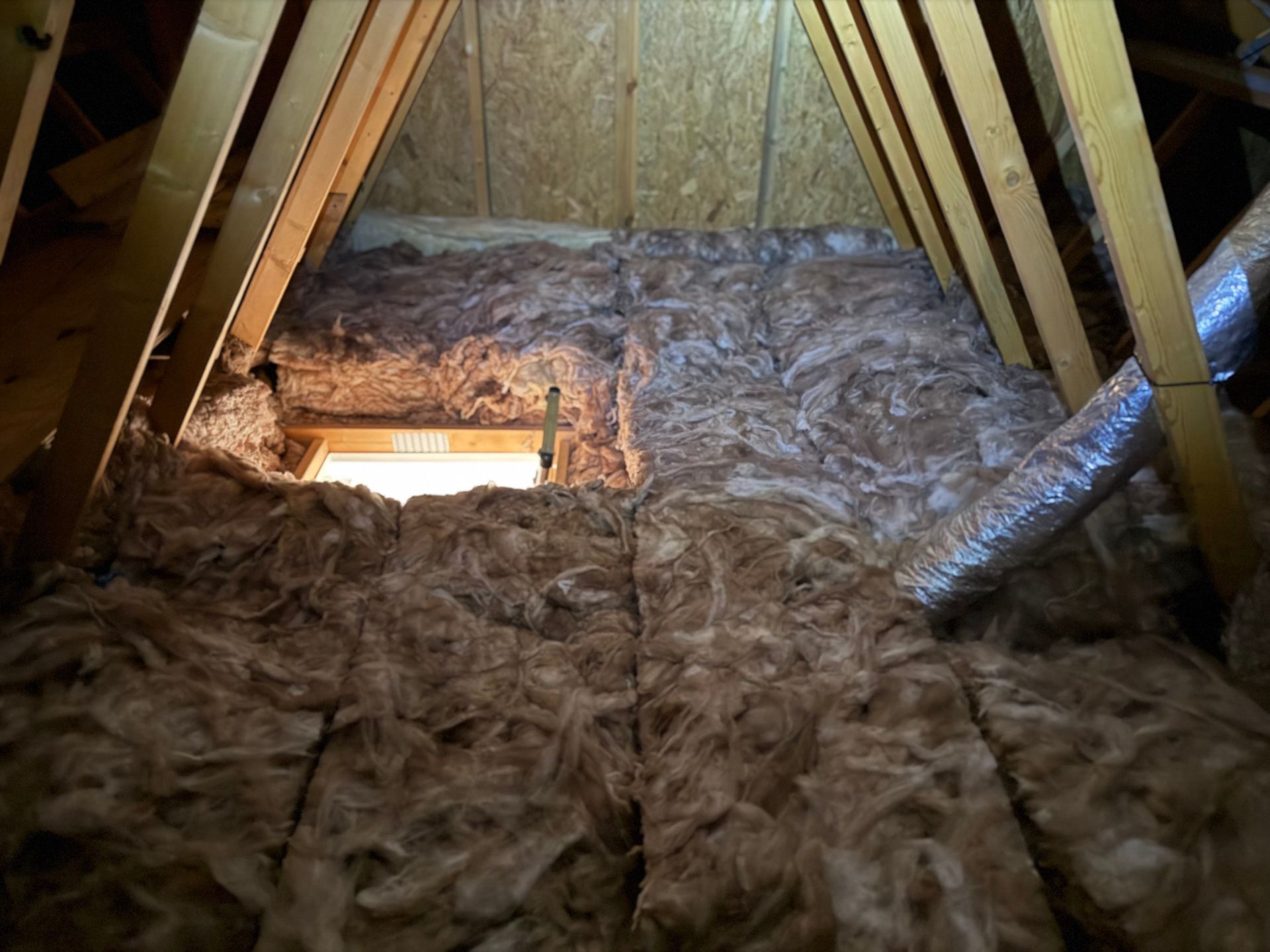 Attic interior with insulation between rafters, a duct, and a small opening, lit from below.