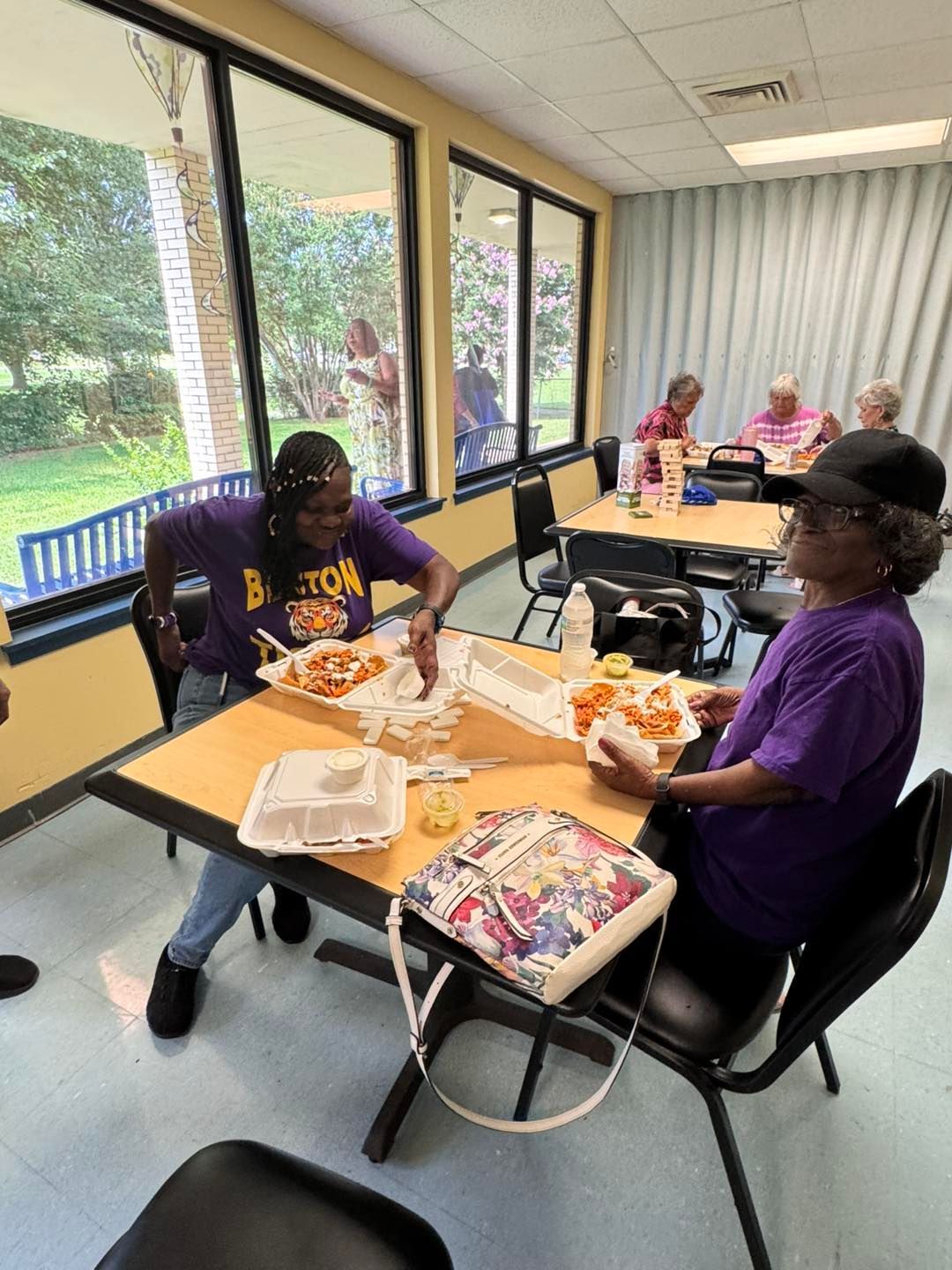 Two women in purple shirts eat at a table indoors. Other people are eating at nearby tables.