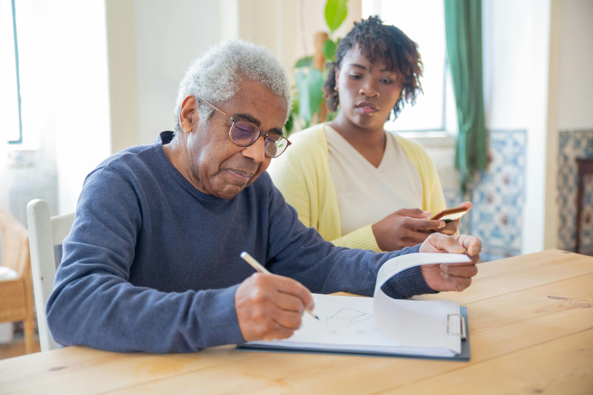 Elderly man signing document at a table, woman looks on holding a phone.