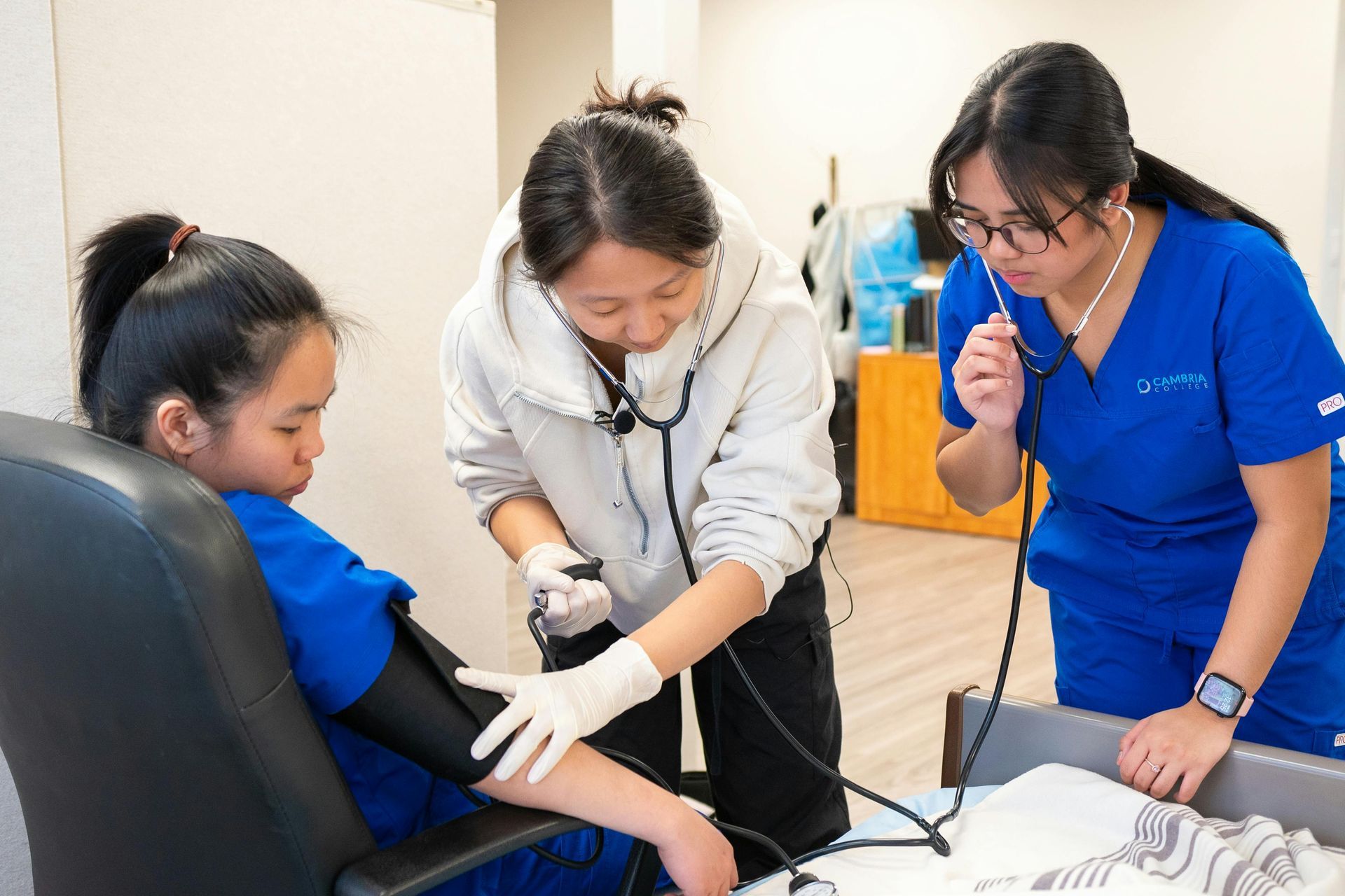 Nurse checking a seated patient's blood pressure while another clinician observes in a clinic room