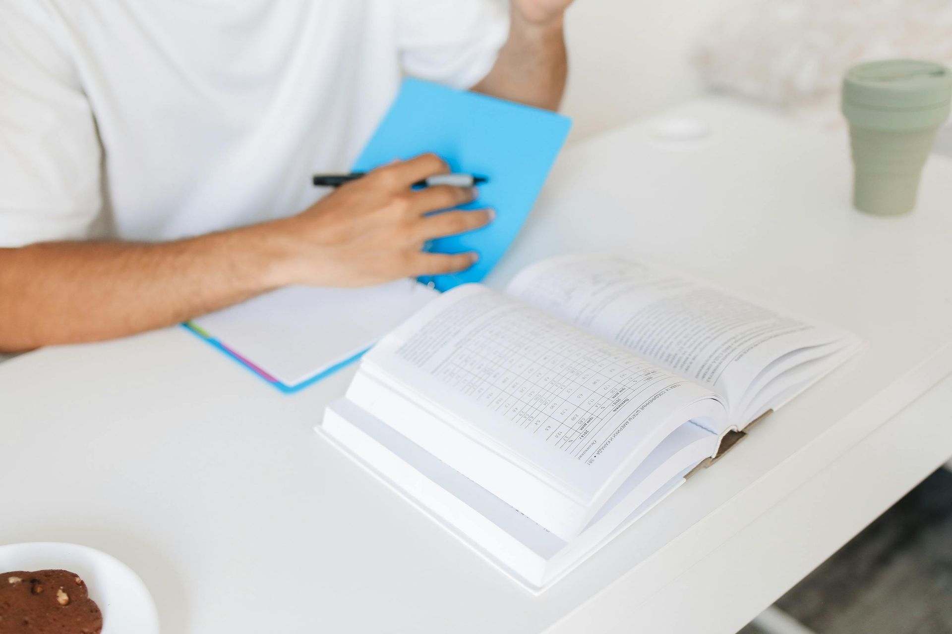 Person writing in a blue notebook beside an open textbook on a white table