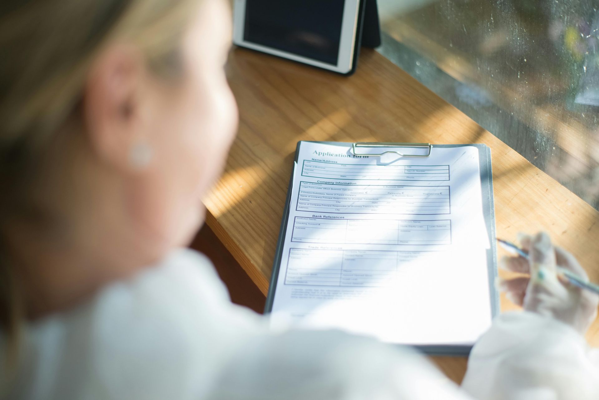 Person writing on a clipboard at a sunlit desk near a window, with a tablet nearby