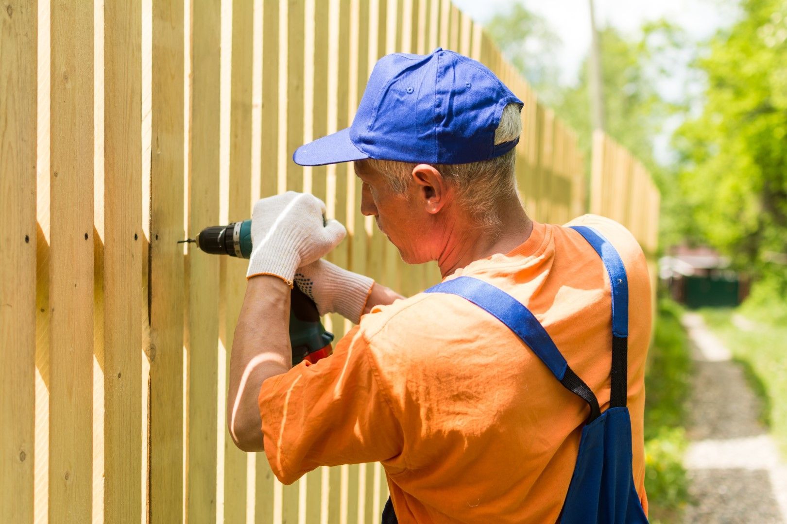 Man in orange shirt and blue cap drilling a wooden fence outdoors.