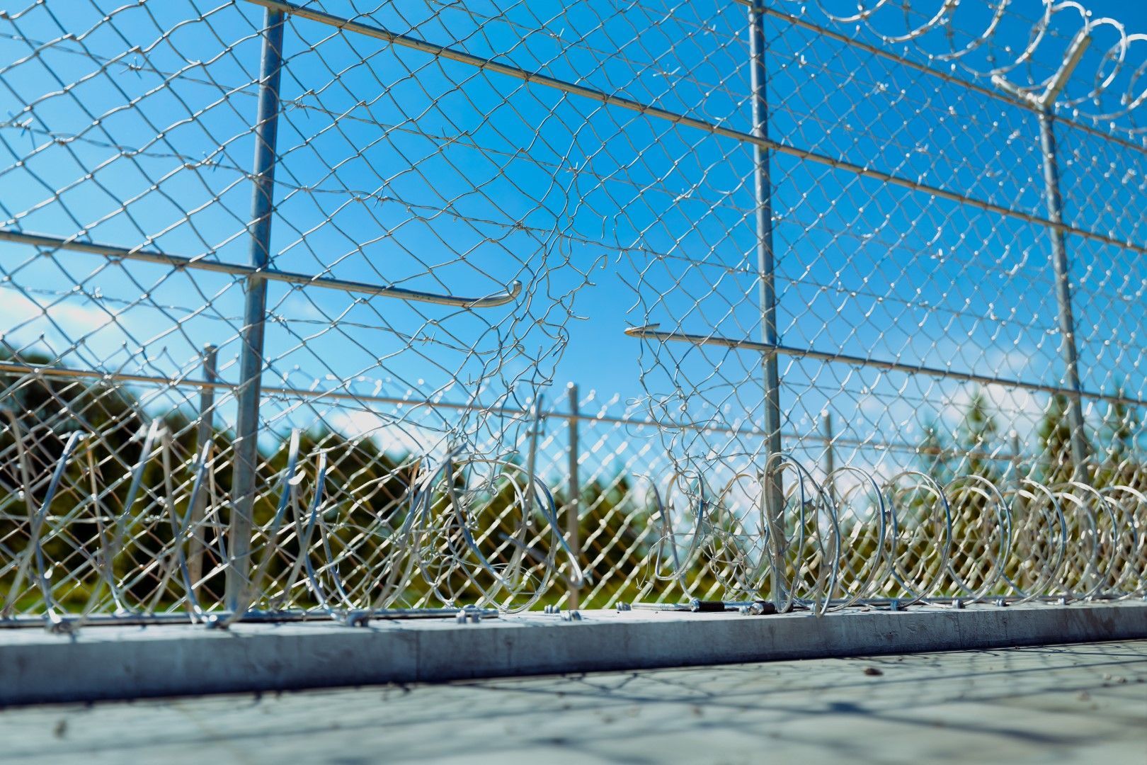 Chain-link fence with razor wire on top against a blue sky. Trees are visible in the background.