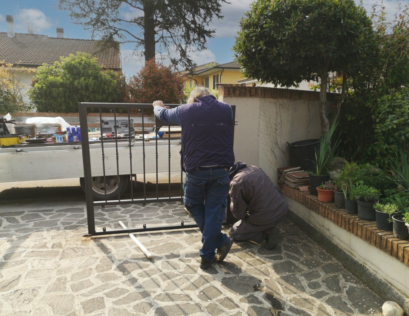 Two men installing a wrought iron gate on a paved driveway next to a brick wall.