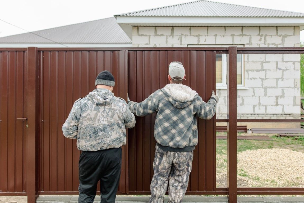 Two people installing a brown metal fence outdoors near a light-colored building.