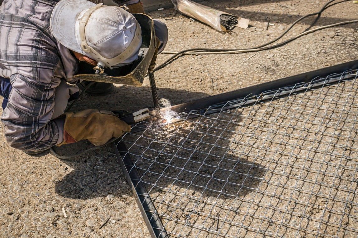 Welder in a plaid shirt and helmet working on a chain-link fence outdoors. Sparks fly.