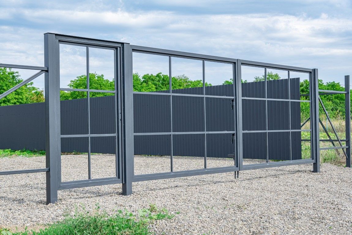 Gray metal gate with glass panels, in front of a dark gray fence and trees.
