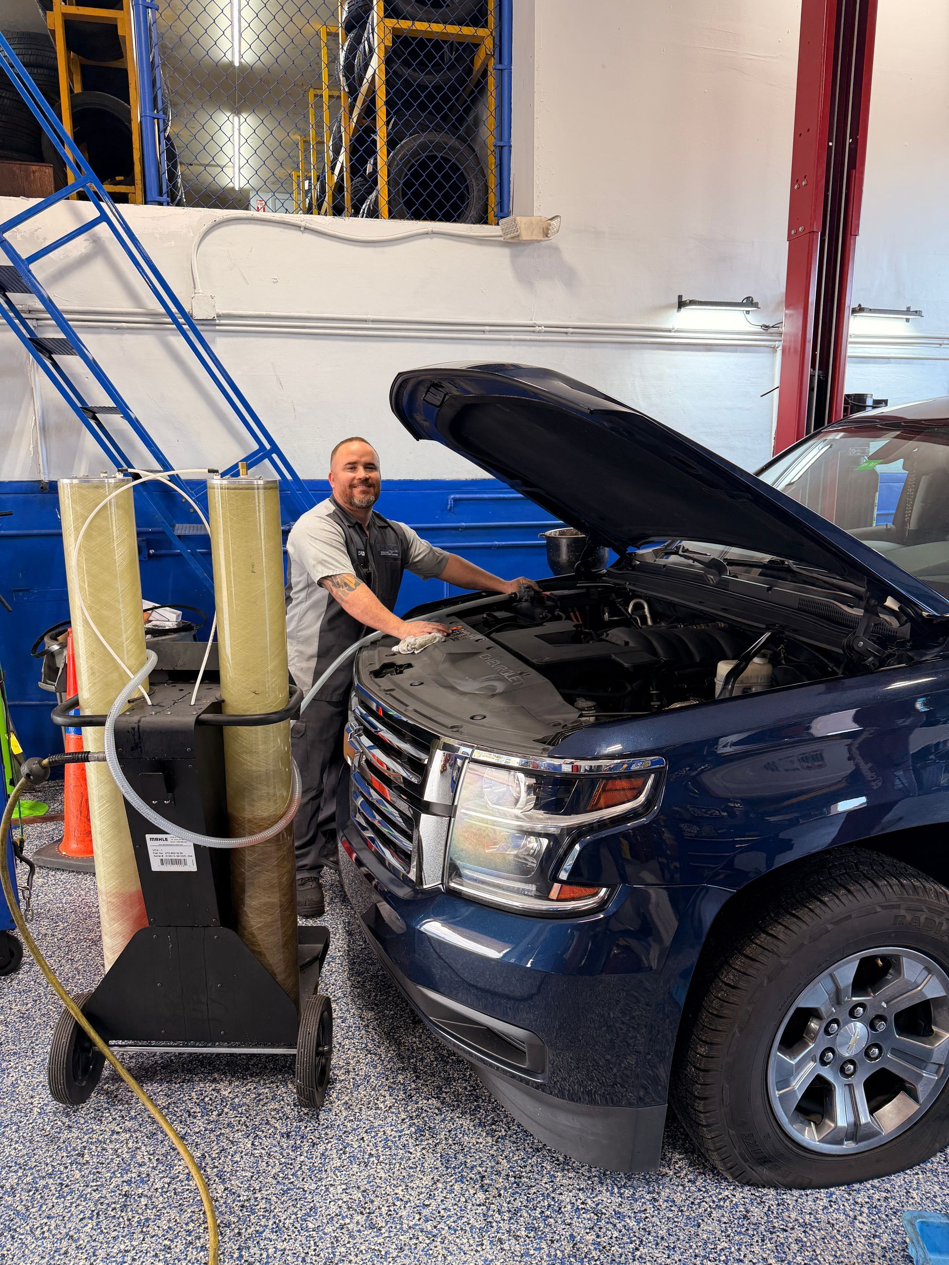 Mechanic working on a blue car engine in a garage. He smiles. A fluid machine is nearby.