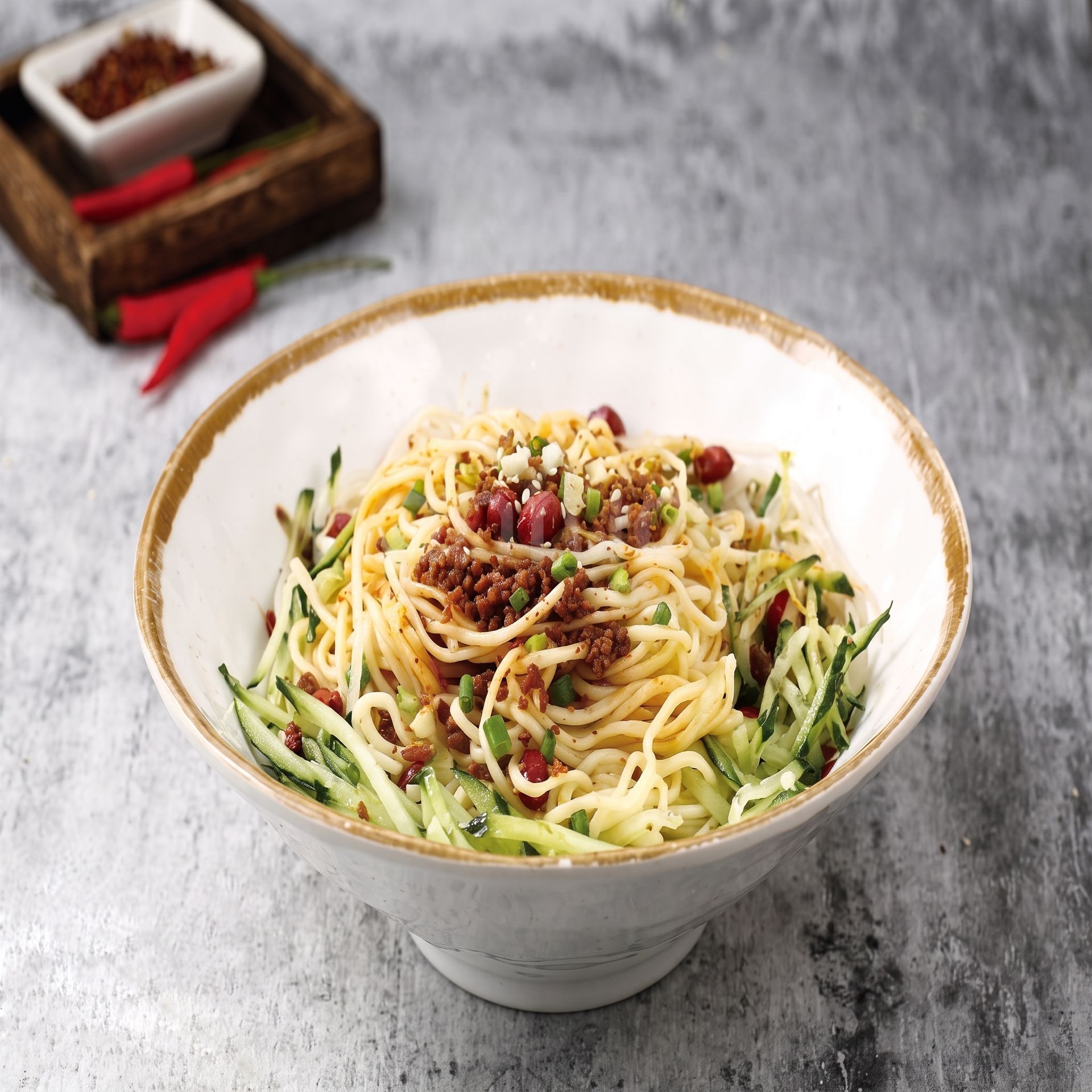 Bowl of spaghetti topped with meat sauce and zucchini, served on a gray table with chili peppers nearby