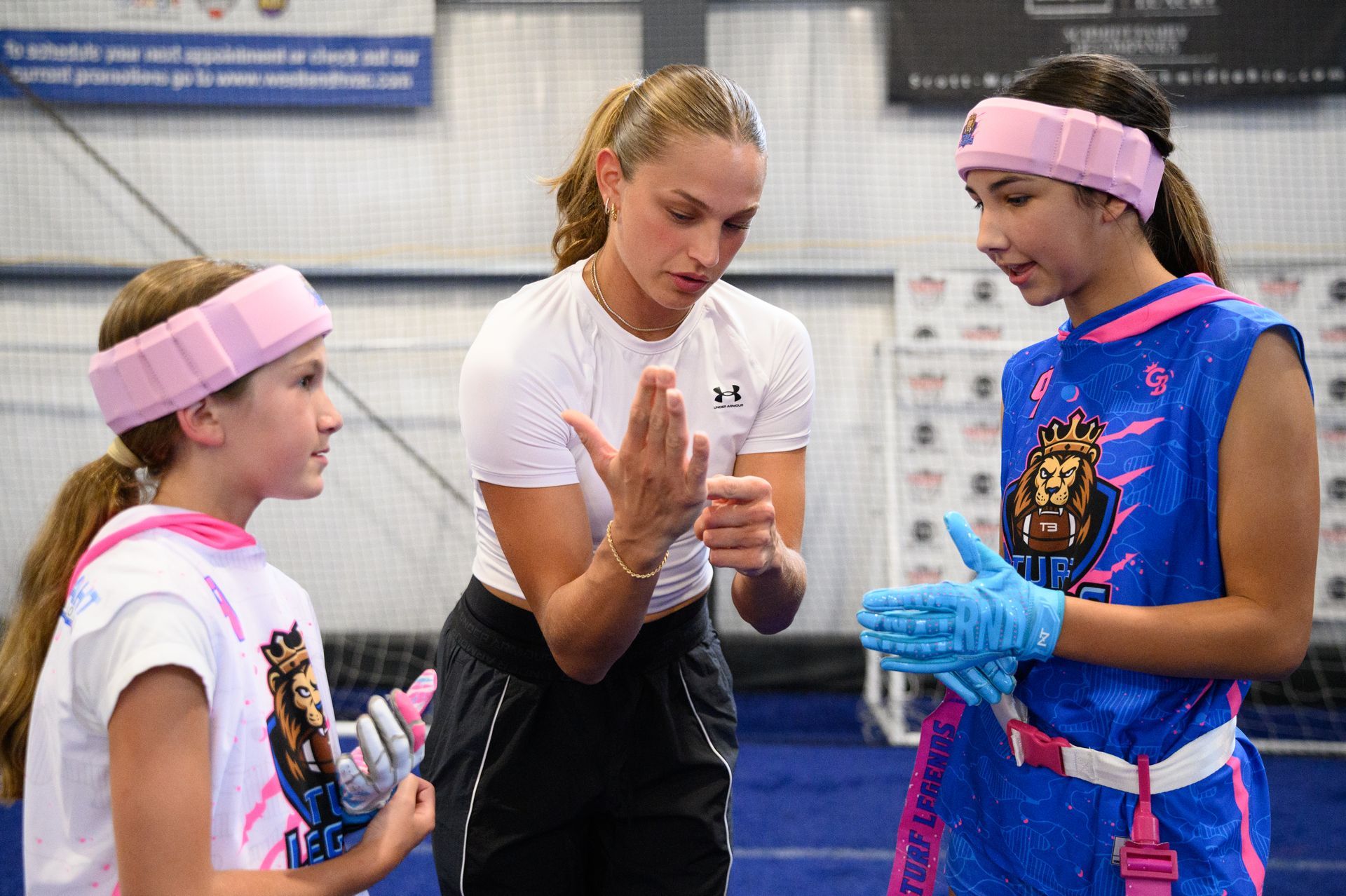 Two people in athletic gear listen to a coach explaining hand positions in an indoor training gym