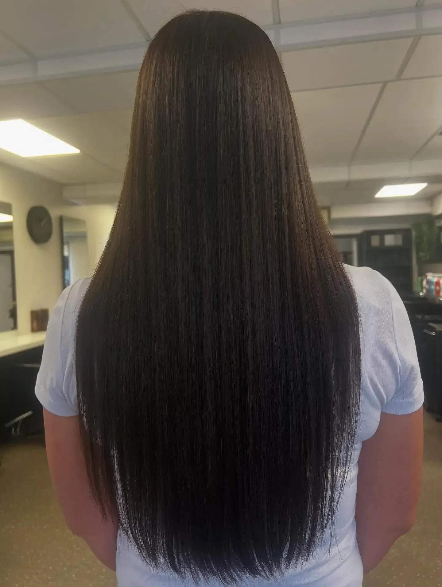 Person with long dark hair standing indoors under fluorescent lights, seen from behind