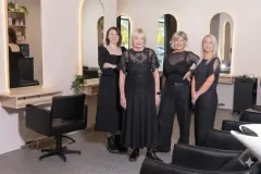 Four women standing in a bright salon, dressed in black, smiling at the camera.