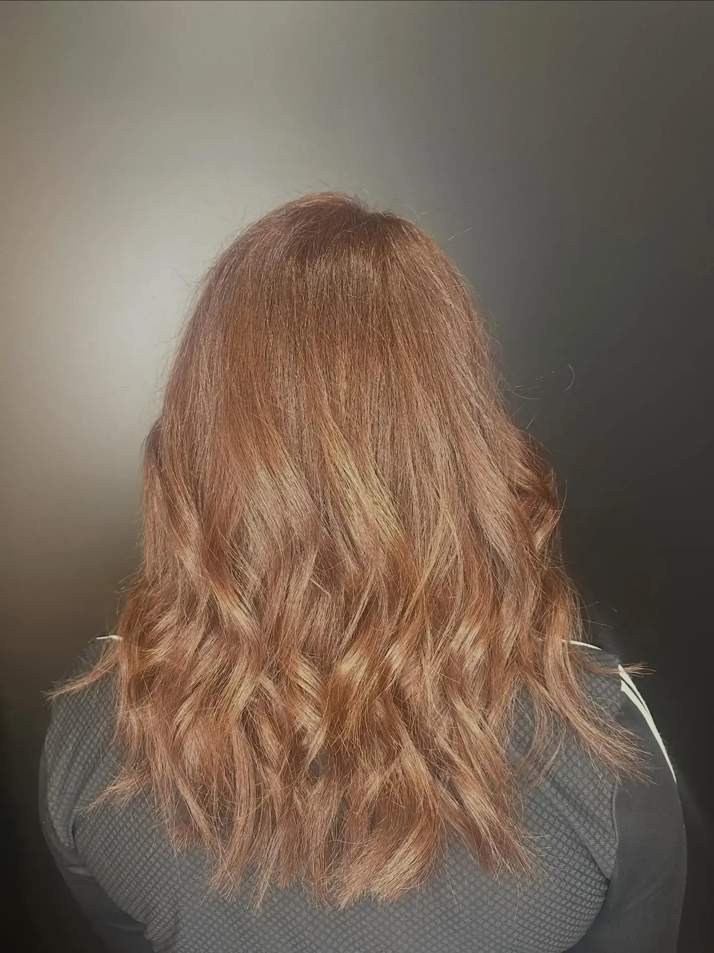 Back view of a person with shoulder-length wavy brown hair against a plain gray wall.