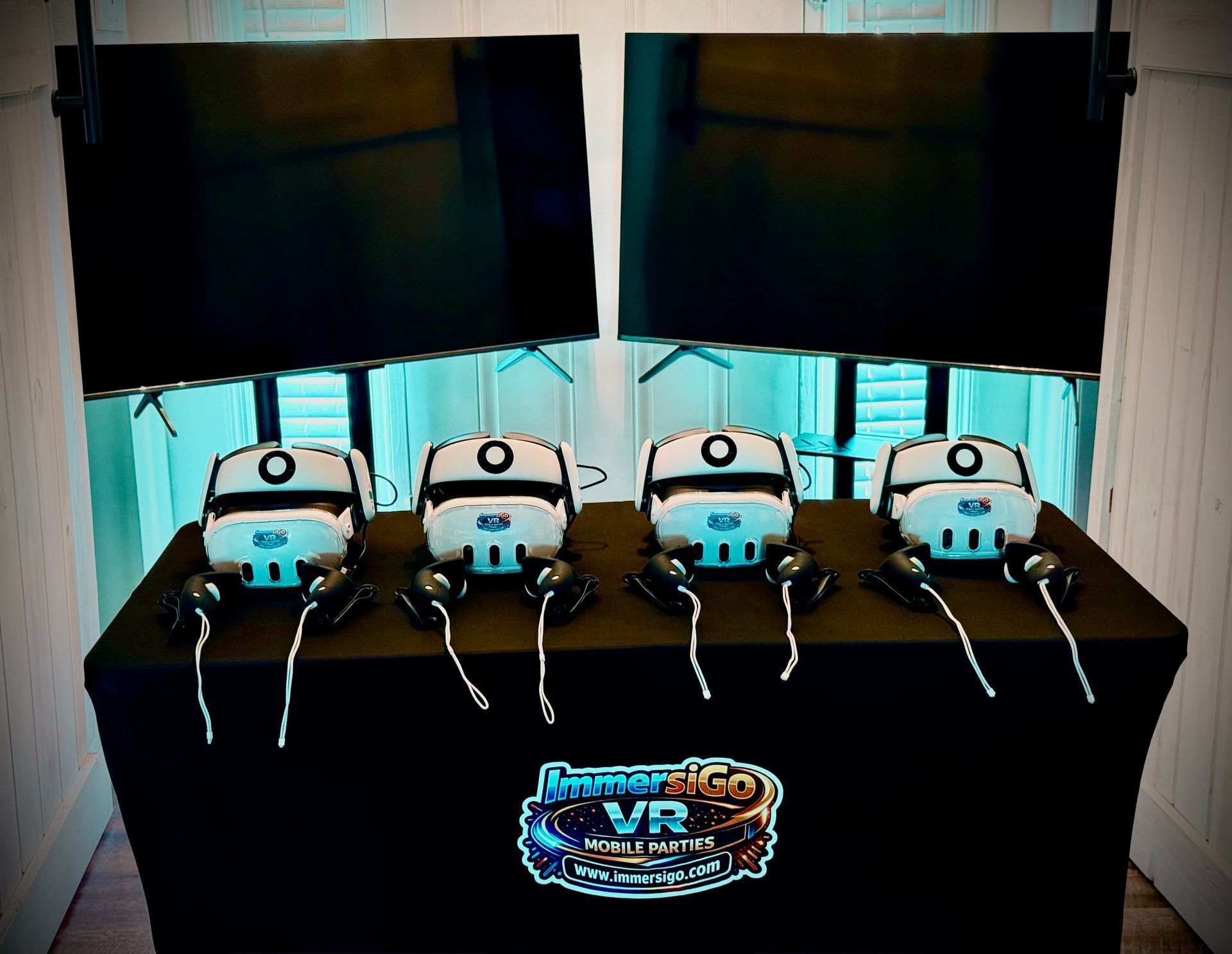 Four VR headsets and controllers on a table beneath two large monitors at a demo booth