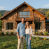 Couple standing in front of a rustic wooden house with mountain views and a green lawn
