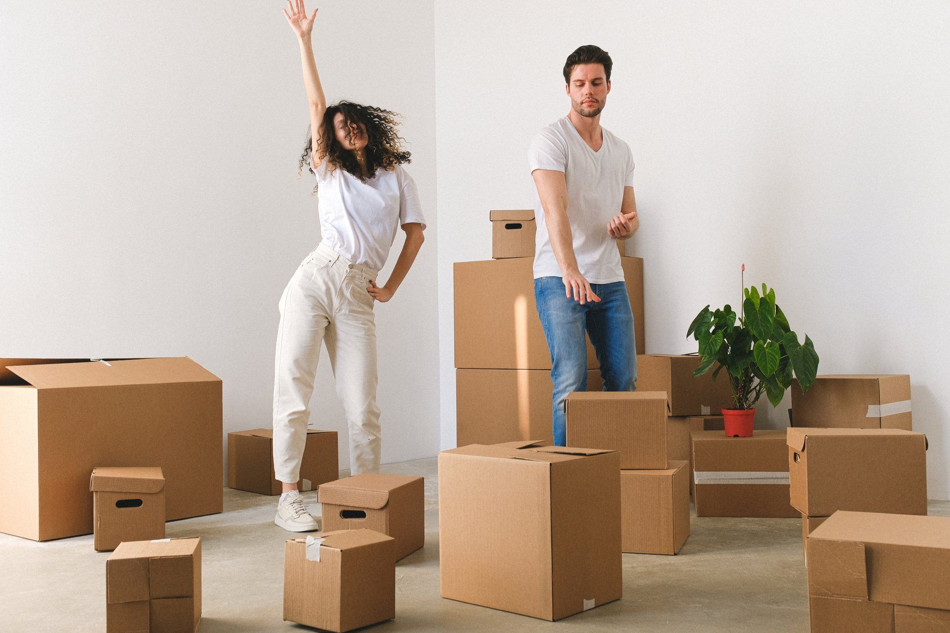 Two people in a sparse room surrounded by moving boxes, one waving and one standing near a plant.