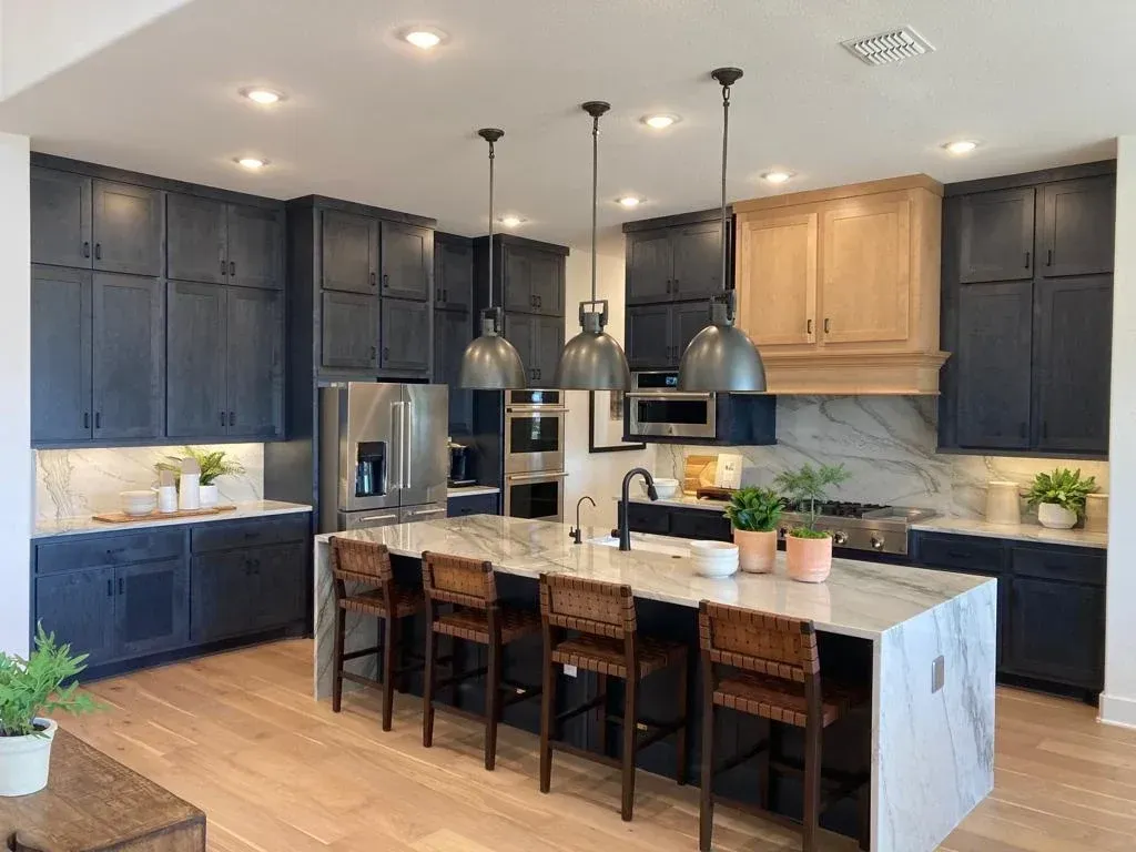 Modern kitchen with dark blue cabinets, light wood floor, island with marble countertop, and pendant lights.