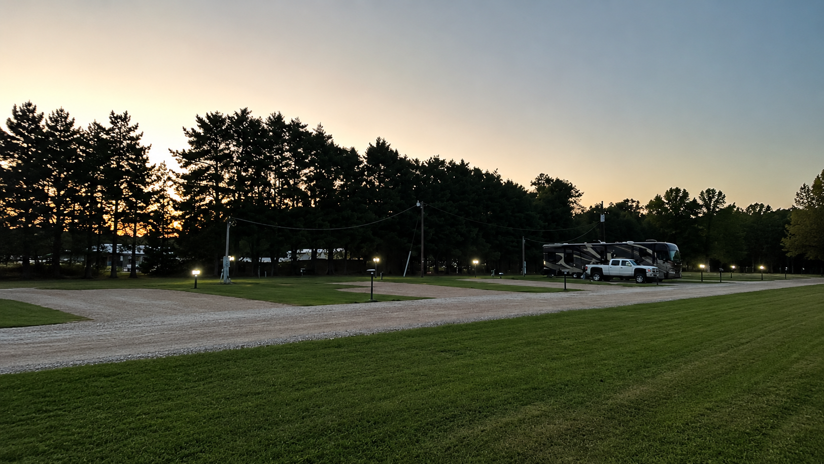 Dusk over a grassy field and gravel drive beside tall trees, with an RV parked near the edge.