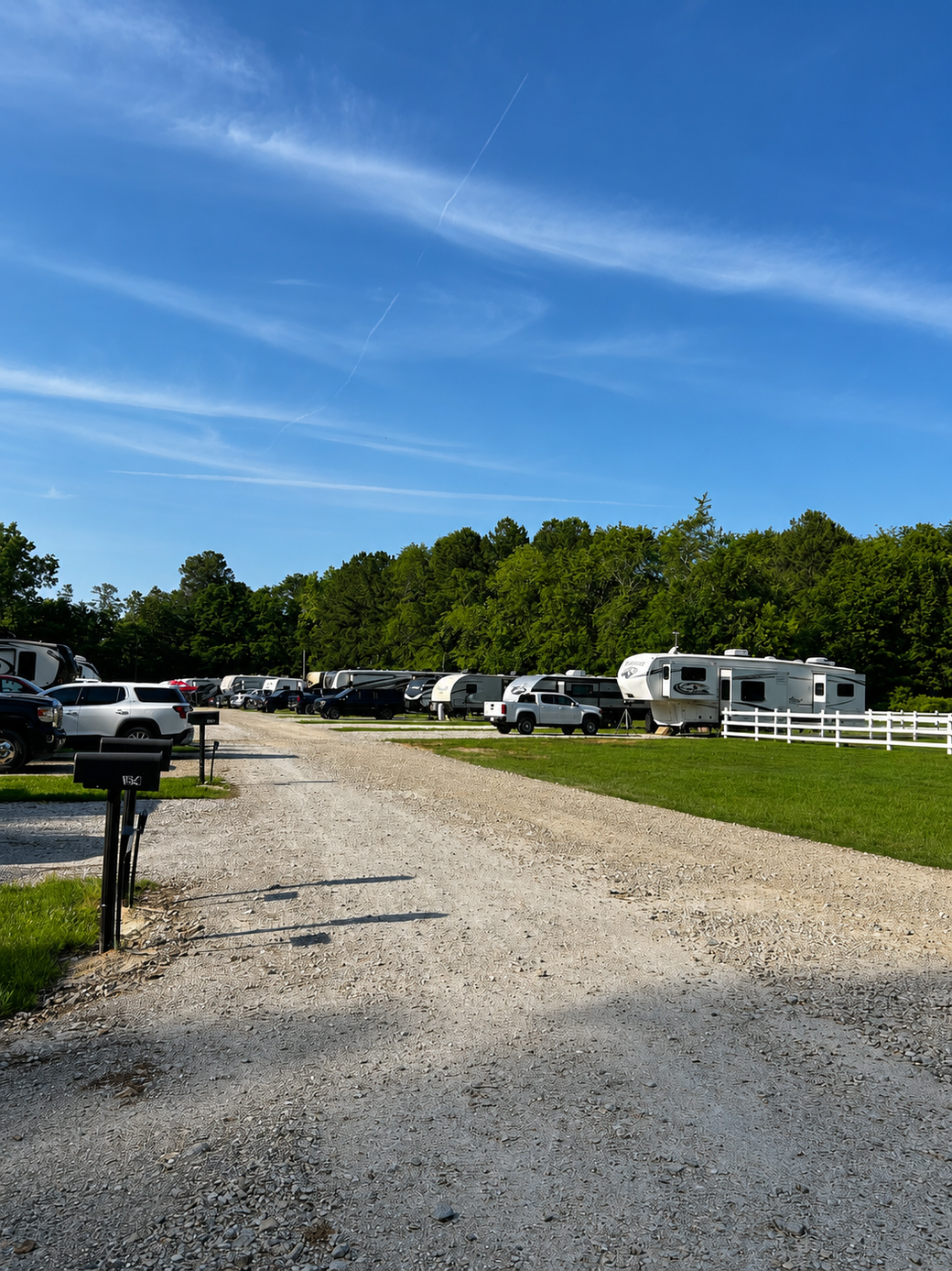 Gravel lot with parked cars and RVs beside a grassy field under a blue sky