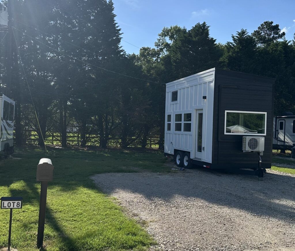 Small gray tiny house on a gravel drive beside trees and grass in bright sunlight