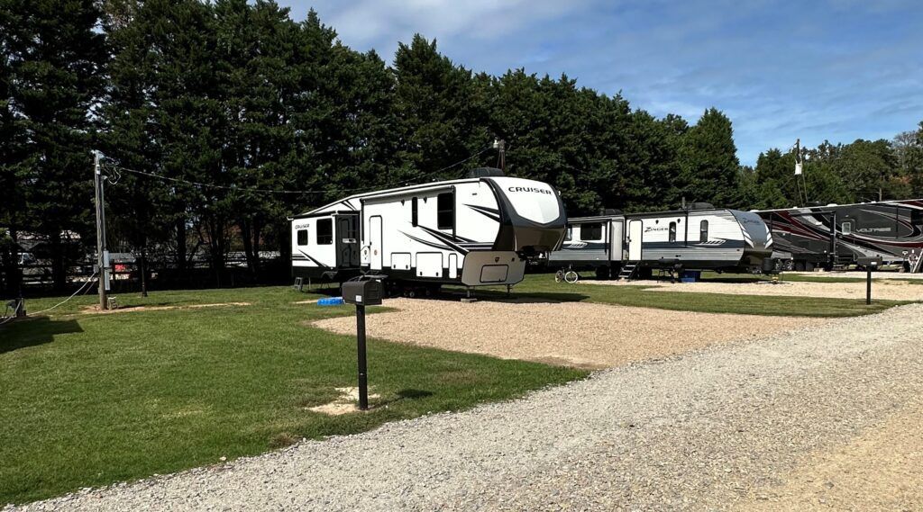 Rv trailers parked on grassy lots beside a gravel road, with trees in the background.