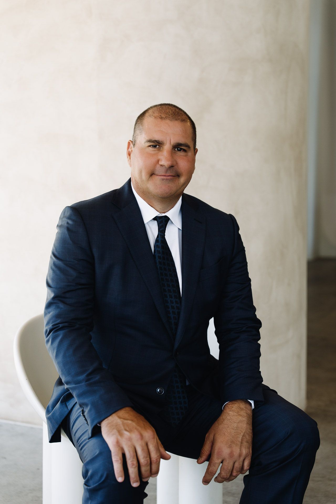 Man in a navy suit sits, smiles, and looks at the camera.