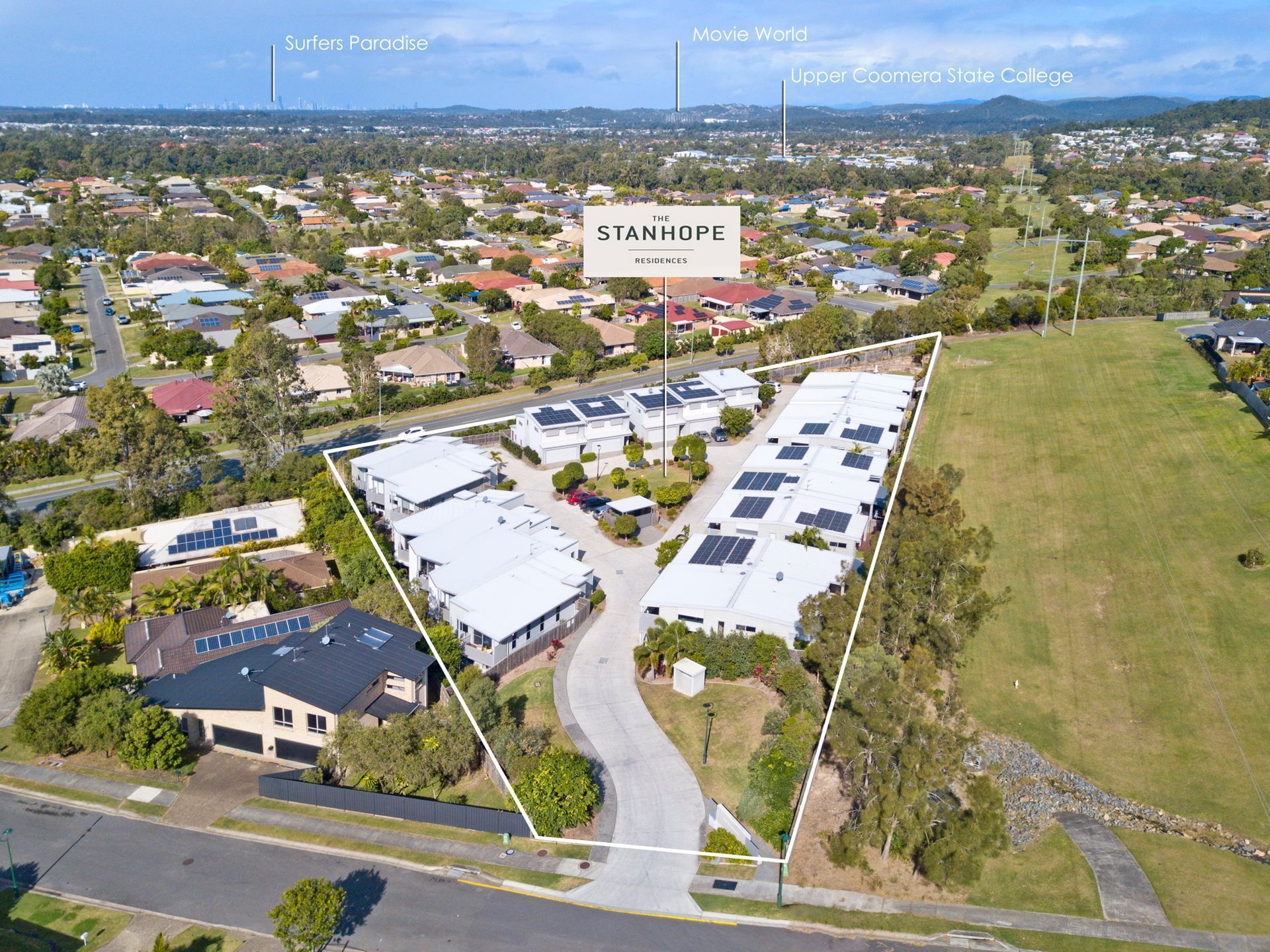 Aerial view of a white-roofed commercial complex beside a large grassy field in a suburban area.
