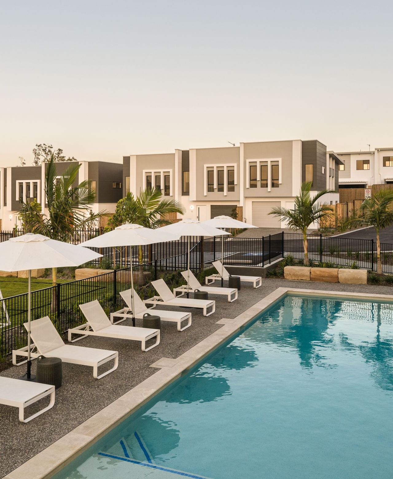 Poolside lounge chairs and umbrellas beside a swimming pool, with modern buildings in the background.