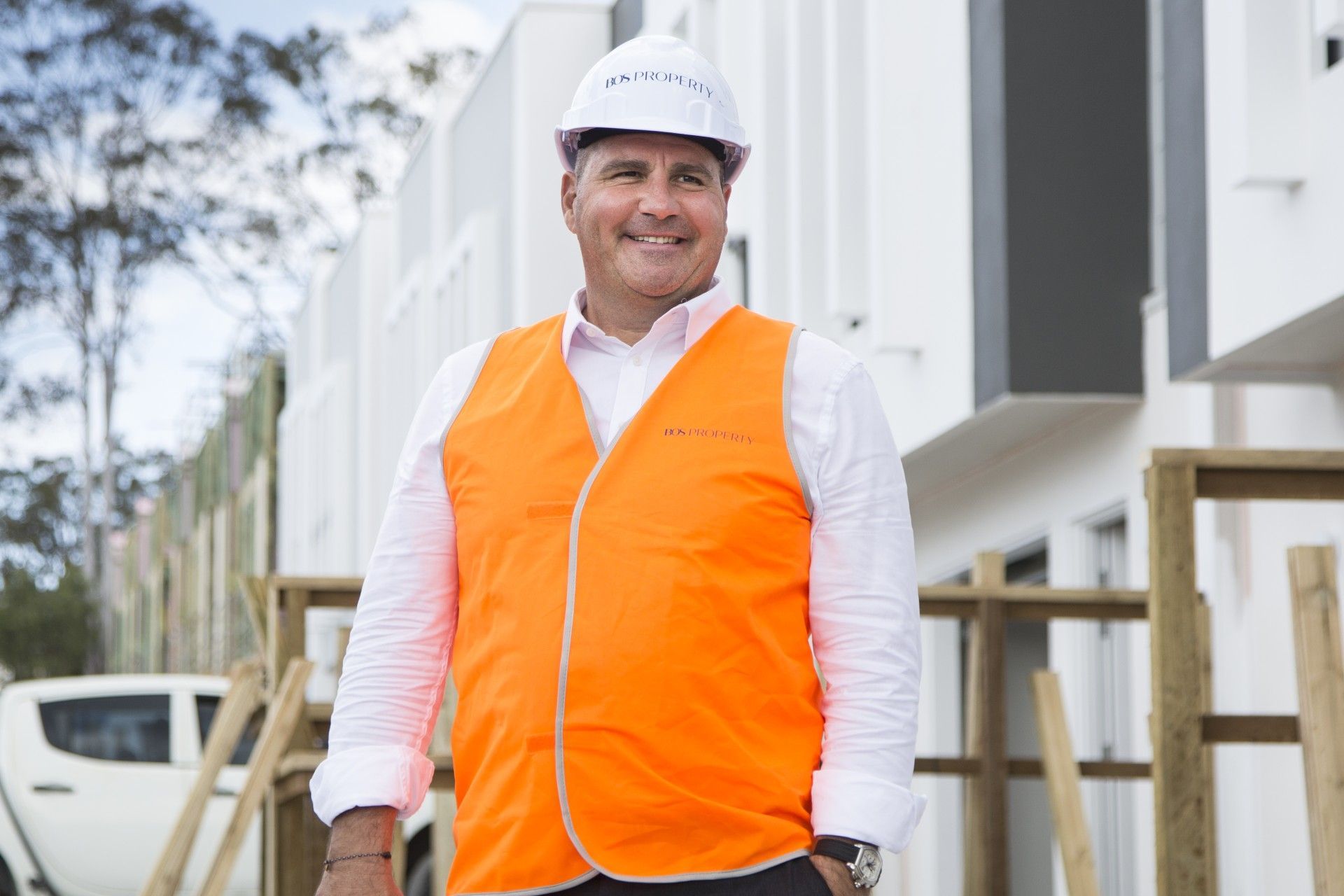 Man in orange vest and hard hat smiles outdoors, construction site in background.
