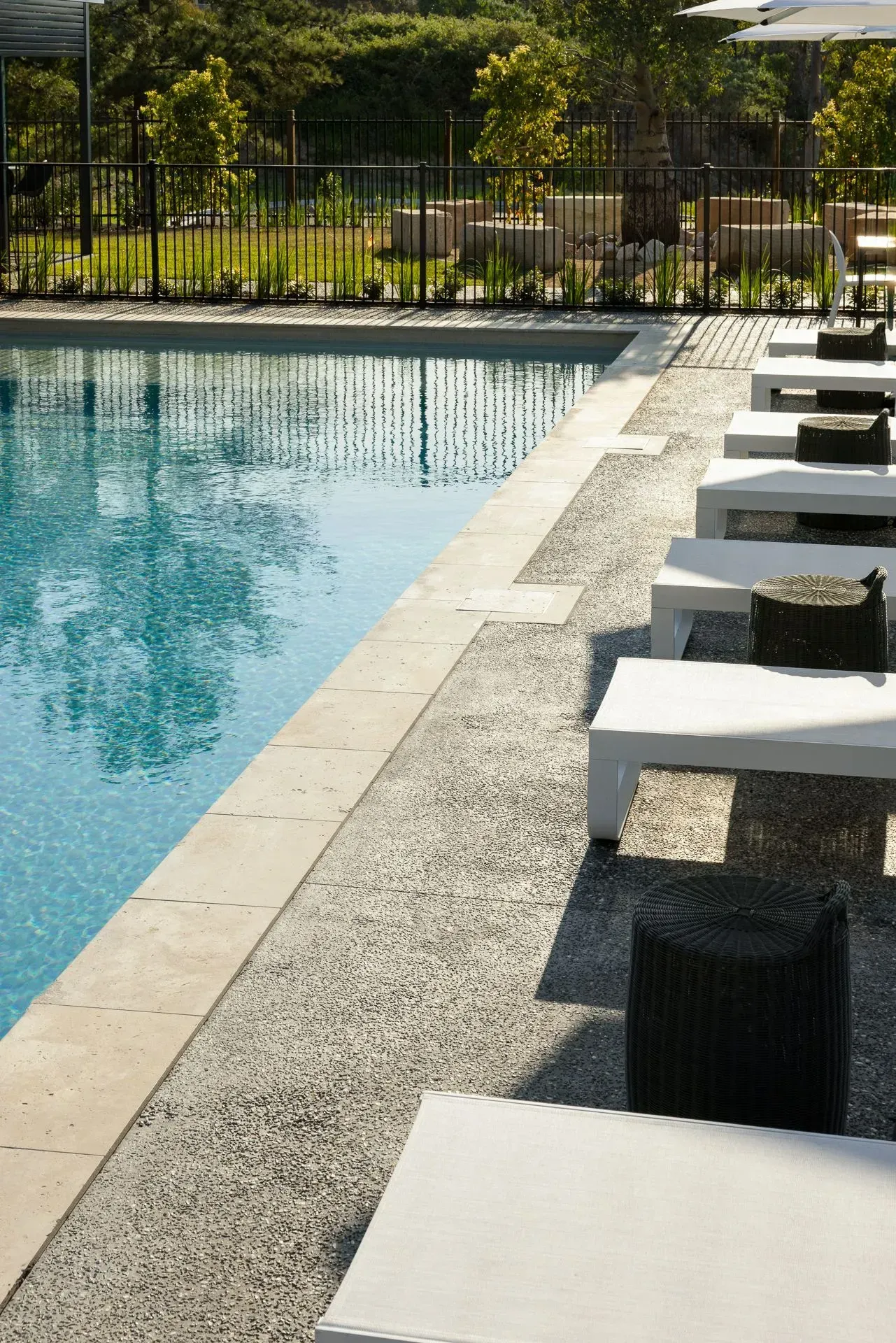 Outdoor swimming pool with white lounge chairs along the deck and a fence with greenery beyond.